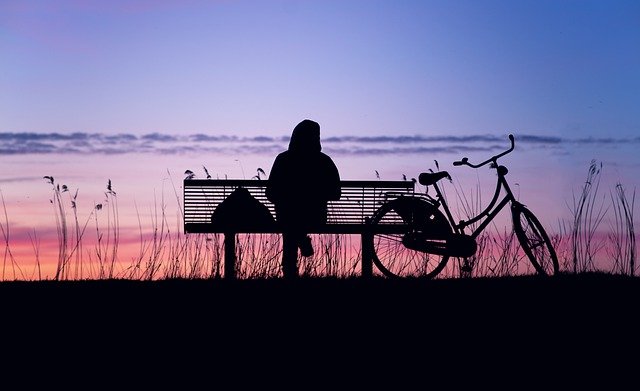 Silhouetted image of person sitting on a bench at sunset, bike beside bench